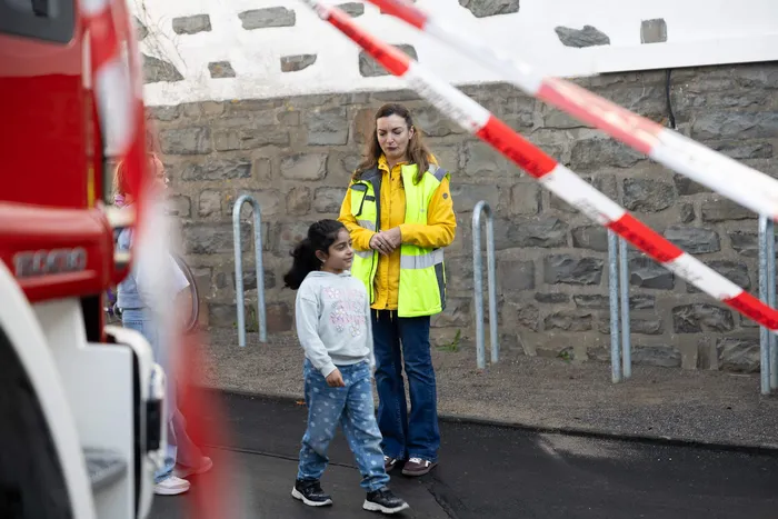 Eine Frau in einer gelben Warnweste steht neben einem kleinen Mädchen vor einer Steinmauer und rot-weißen Absperrungen.