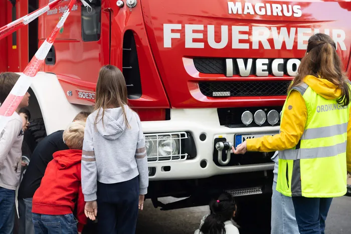Kinder und eine Person in gelber Warnweste stehen vor einem roten Feuerwehrfahrzeug bei einer Übung zum toten Winkel.