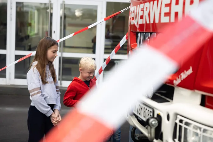 Zwei Kinder stehen hinter rot-weißem Absperrband und betrachten ein Feuerwehrauto während einer Übung zum toten Winkel.