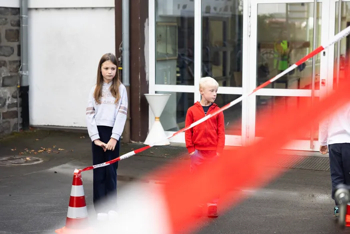 Zwei Kinder stehen hinter einem rot-weißen Absperrband während einer Übung zum Thema „Toter Winkel“ vor einem Gebäude.