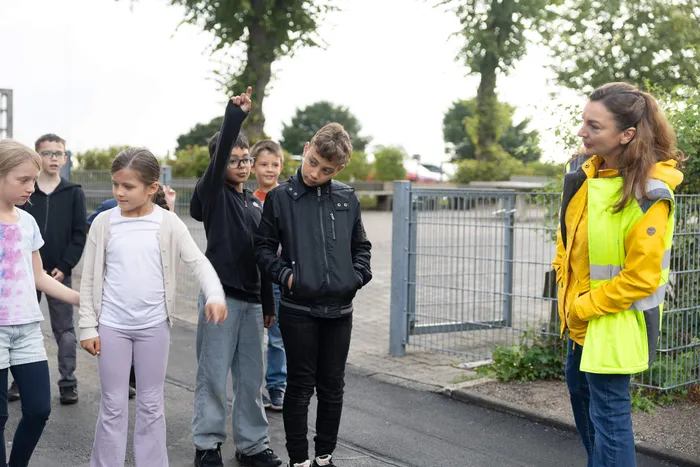 Eine Gruppe Grundschüler bei einer praktischen Übung im Freien, ein Junge zeigt nach oben, während eine Frau in einer gelben Warnweste die Kinder beobachtet.