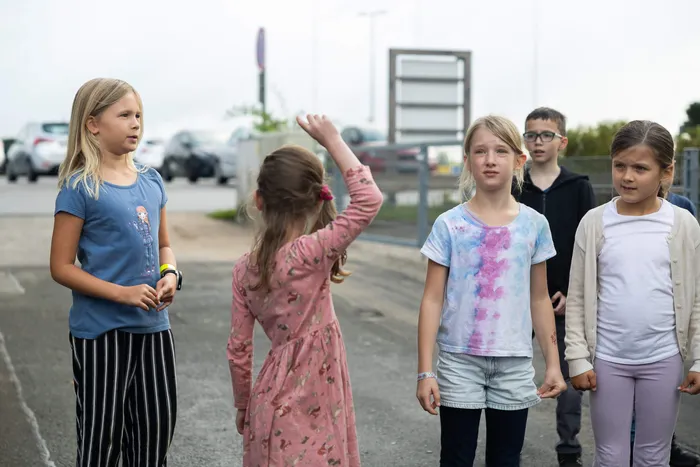 Vier Grundschulkinder stehen auf einem asphaltierten Platz, ein Mädchen in einem rosa Kleid hebt den Arm.