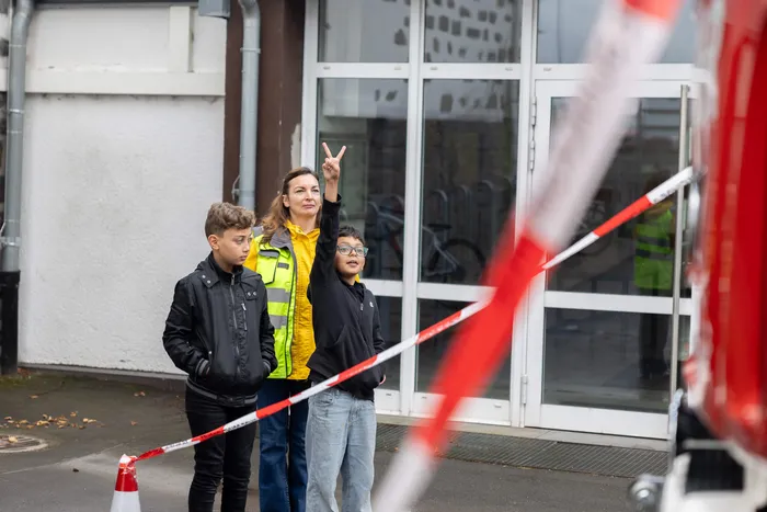 Eine Frau in einer gelben Warnweste steht mit zwei Jungen hinter einem rot-weißen Absperrband; einer der Jungen zeigt das Peace-Zeichen.
