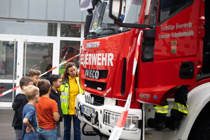 Eine Frau in einer gelben Warnweste spricht mit einer Gruppe von Kindern vor einem roten Feuerwehrfahrzeug.
