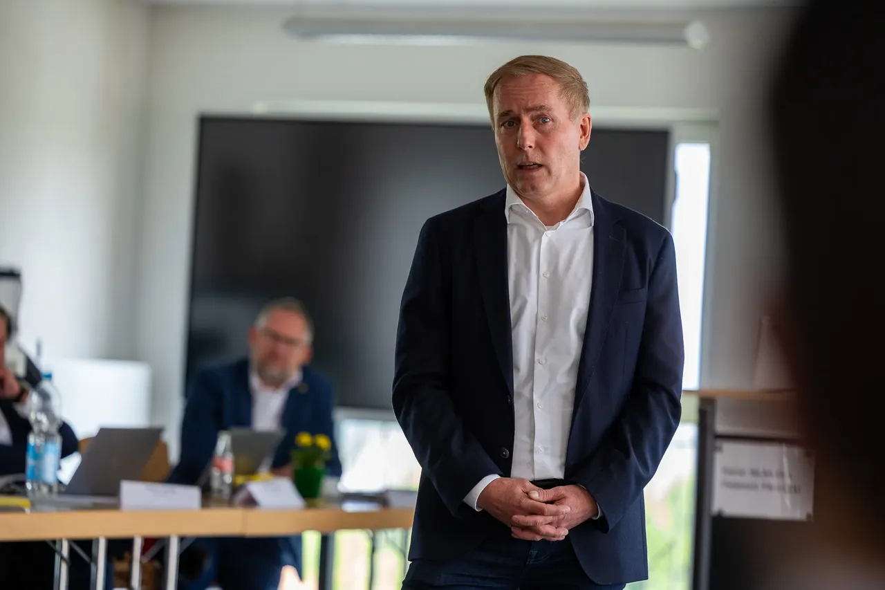 Ein Mann in einem dunklen Anzug und offenem Hemd spricht in einem Konferenzraum. Im Hintergrund sitzt ein weiterer Mann an einem Tisch mit Laptops und Unterlagen.
