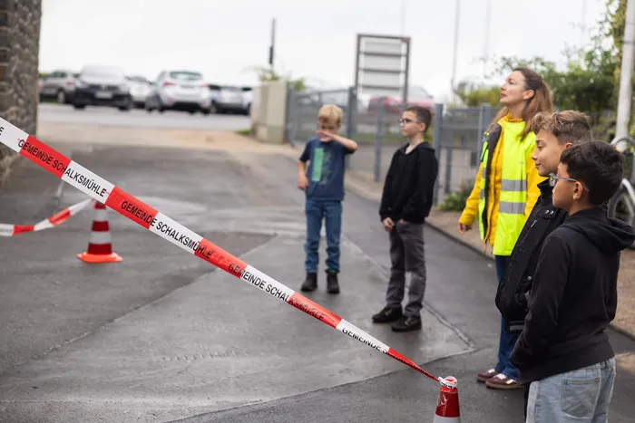 Kinder und eine Frau in einer gelben Warnweste stehen hinter einem rot-weißen Absperrband auf einem asphaltierten Platz.
