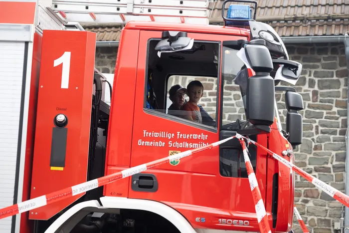 Ein Kind und ein Mann sitzen im Fahrerhaus eines roten Feuerwehrautos der Freiwilligen Feuerwehr Schalksmühle hinter rot-weißem Absperrband.