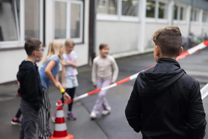 Ein Junge blickt auf eine Gruppe von Kindern hinter einem rot-weißen Absperrband und einem Verkehrskegel bei einer Übung zum „Toten Winkel“.