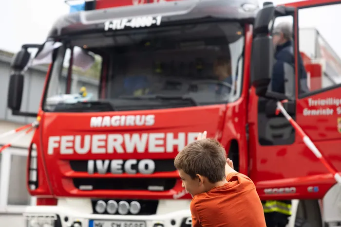 Ein Junge in einem orangefarbenen T-Shirt steht vor einem großen roten Magirus-Feuerwehrauto.