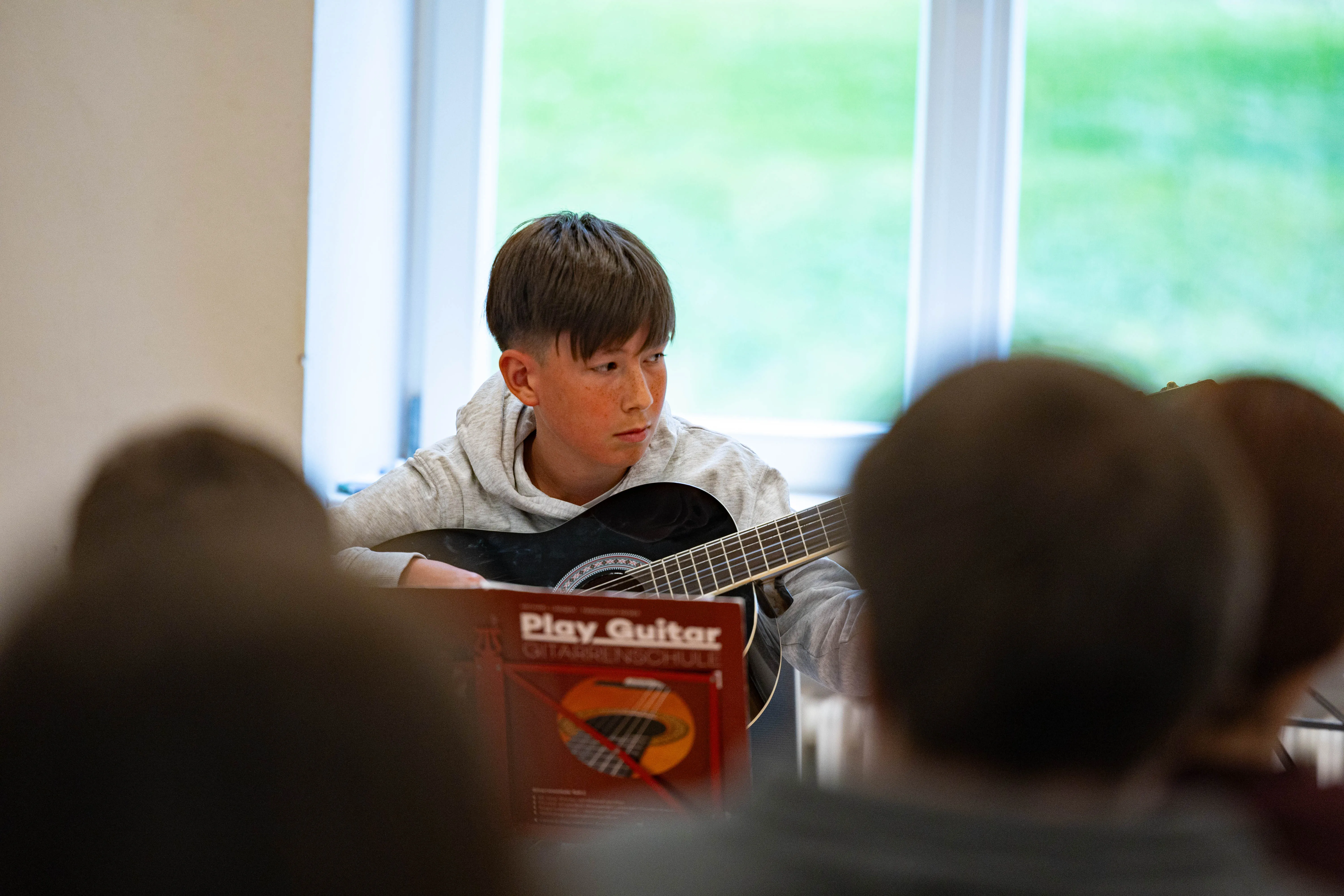 Nuri Sijamic spielt Gitarre während einer Unterrichtsstunde der Musikschule Volmetal. Vor ihm liegt ein Notenbuch mit dem Titel „Play Guitar Gitarrenschule“.