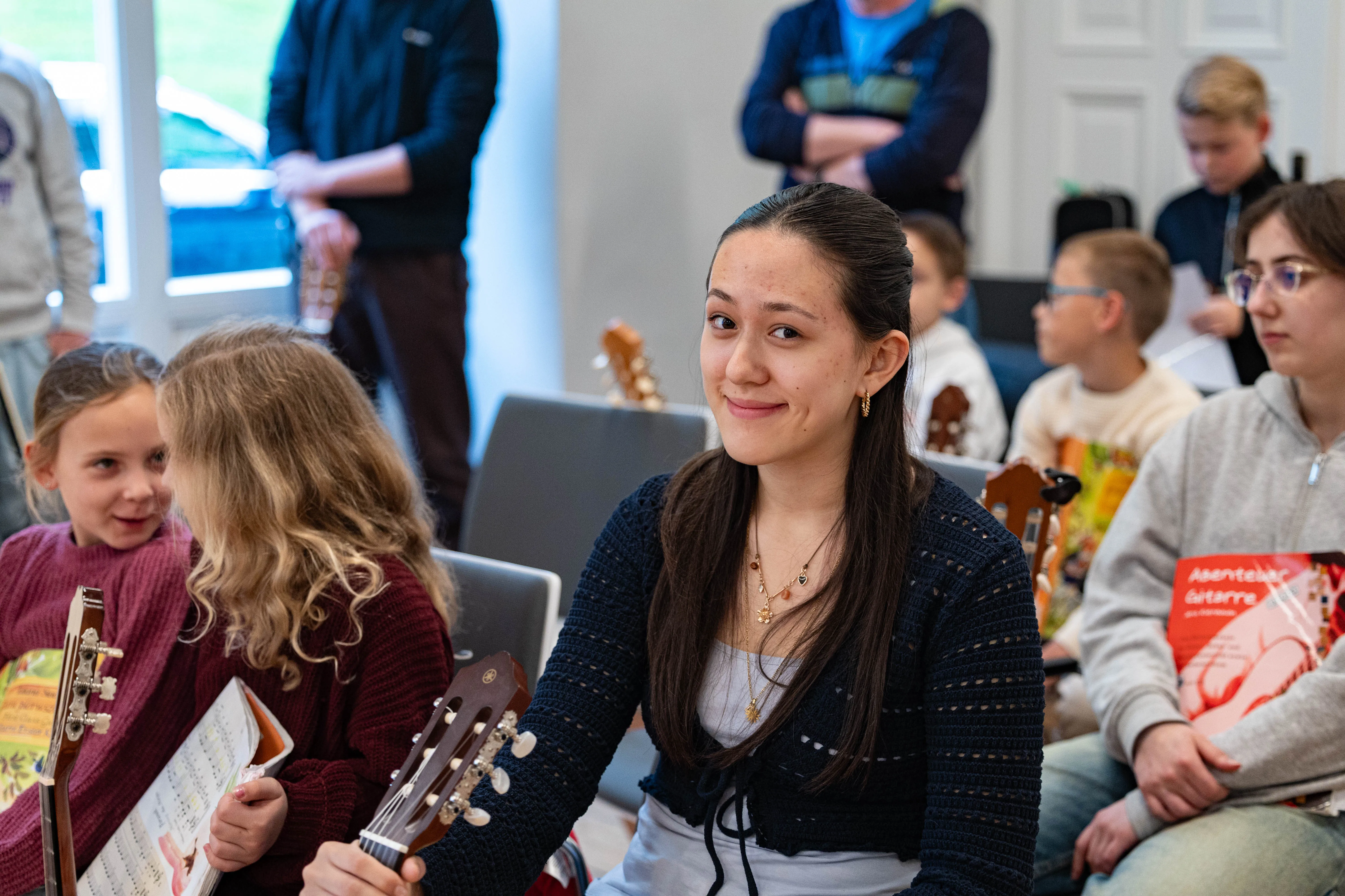 Junge Frau lächelt in einem Raum mit Kindern, die Gitarren halten und Notenblätter betrachten. Im Hintergrund sind weitere Personen und Instrumente zu sehen.