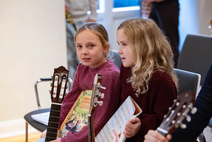 Zwei Mädchen mit Gitarren sitzen auf Stühlen und schauen auf Notenblätter. Im Hintergrund weitere Personen und Musikinstrumente in einem hellen Raum.