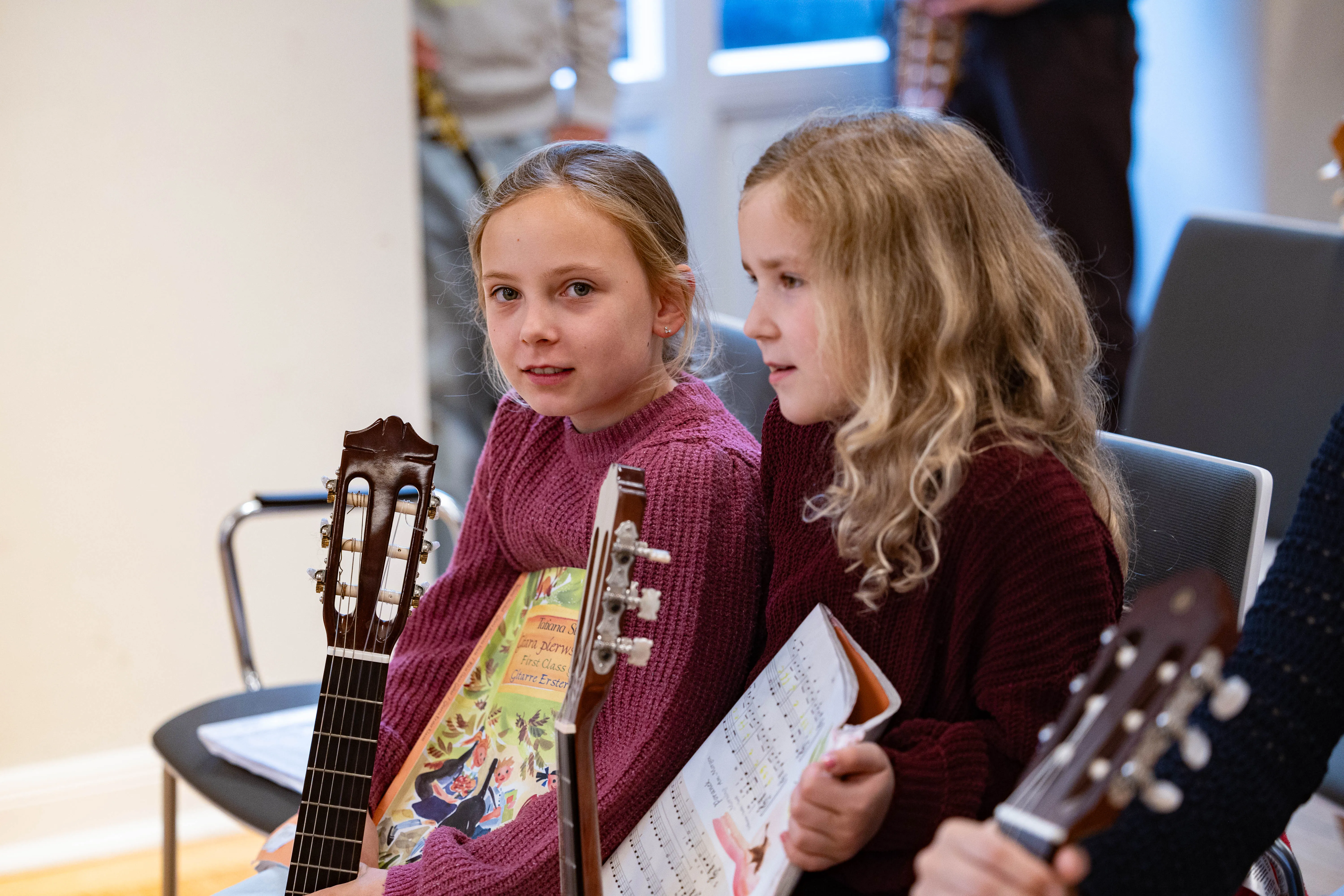 Zwei Mädchen mit Gitarren sitzen auf Stühlen und schauen auf Notenblätter. Im Hintergrund weitere Personen und Musikinstrumente in einem hellen Raum.