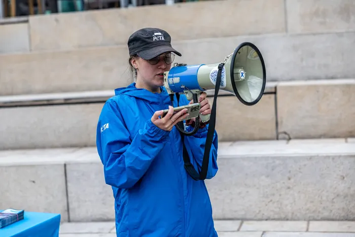 Eine Frau mit PETA-Kappe und blauer Jacke hält ein Megafon und ein Smartphone vor Steinstufen.