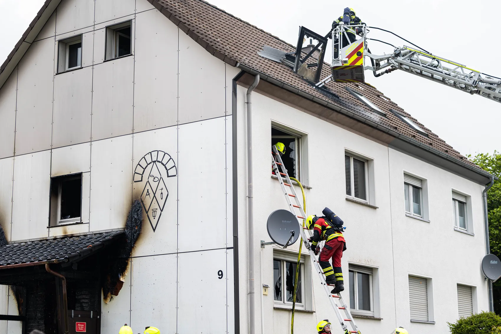 Feuerwehrleute besteigen eine Leiter an einem mehrstöckigen Gebäude mit sichtbaren Brandschäden an der Fassade.