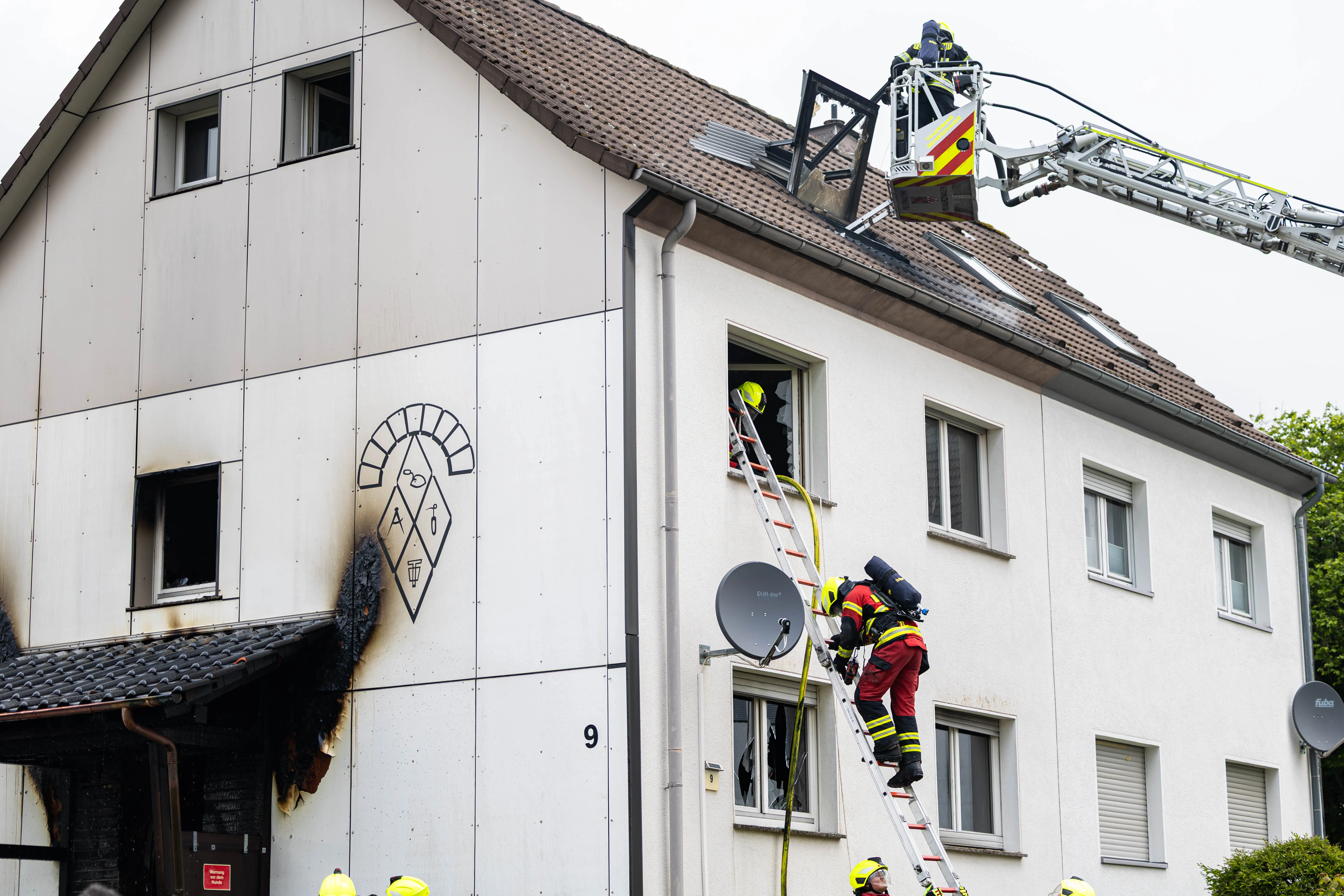 Feuerwehrleute besteigen eine Leiter an einem mehrstöckigen Gebäude mit sichtbaren Brandschäden an der Fassade.