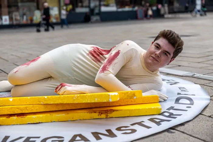Eine Person in einem beigefarbenen, rot gefleckten Body liegt auf einem Banner mit dem Schriftzug „VEGANSTART.DE“ neben gelben Brettern auf dem Boden.
