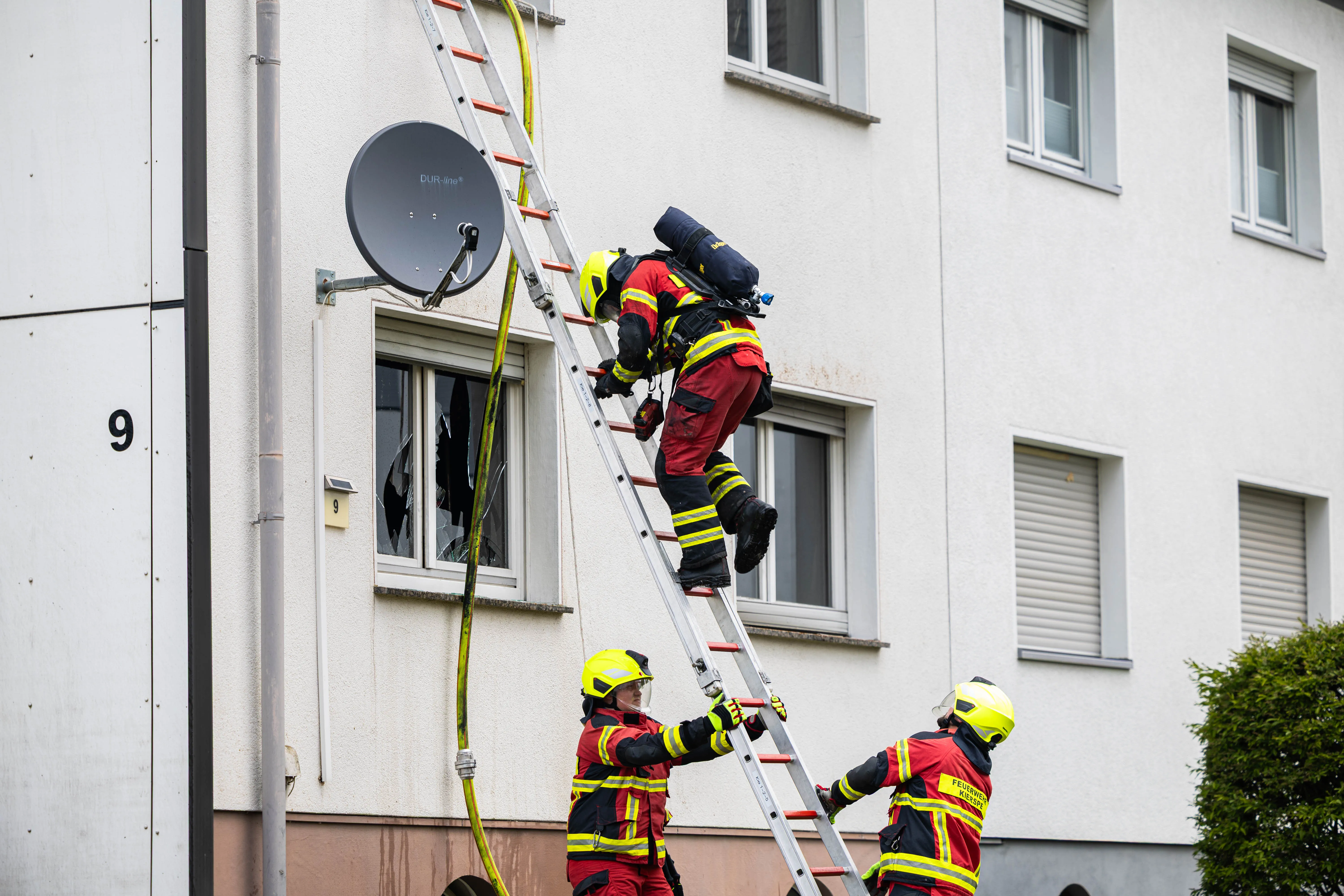 Feuerwehrleute steigen mit einer Leiter an die Fassade eines mehrstöckigen Wohngebäudes, dessen Fenster eingeschlagen sind. Ein Feuerwehrmann klettert gerade hinein.
