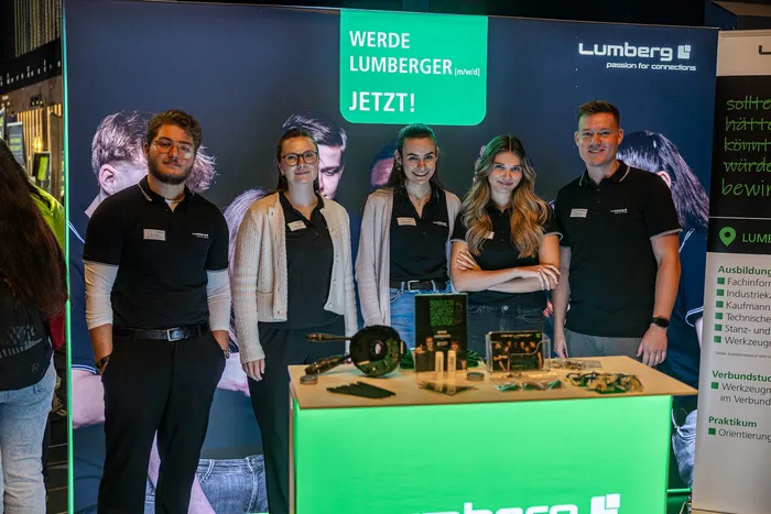 Fünf junge Menschen stehen hinter einem Infostand der Firma Lumberg vor einem Banner mit der Aufschrift „WERDE LUMBERGER JETZT!“.