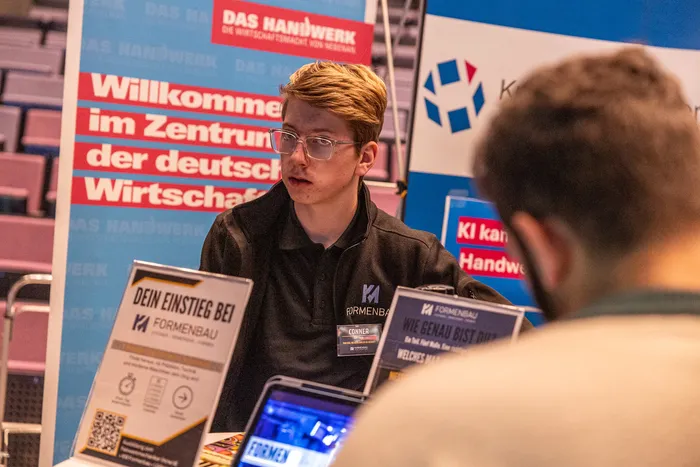 Ein junger Mann im Poloshirt der Firma Formenbau steht an einem Infostand auf einer Messe und spricht mit einer Person im Vordergrund.
