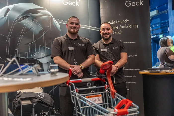 Zwei lächelnde Männer in schwarzen Poloshirts mit dem „Geck“-Logo stehen an einem Messestand vor einem Banner für Ausbildungen.