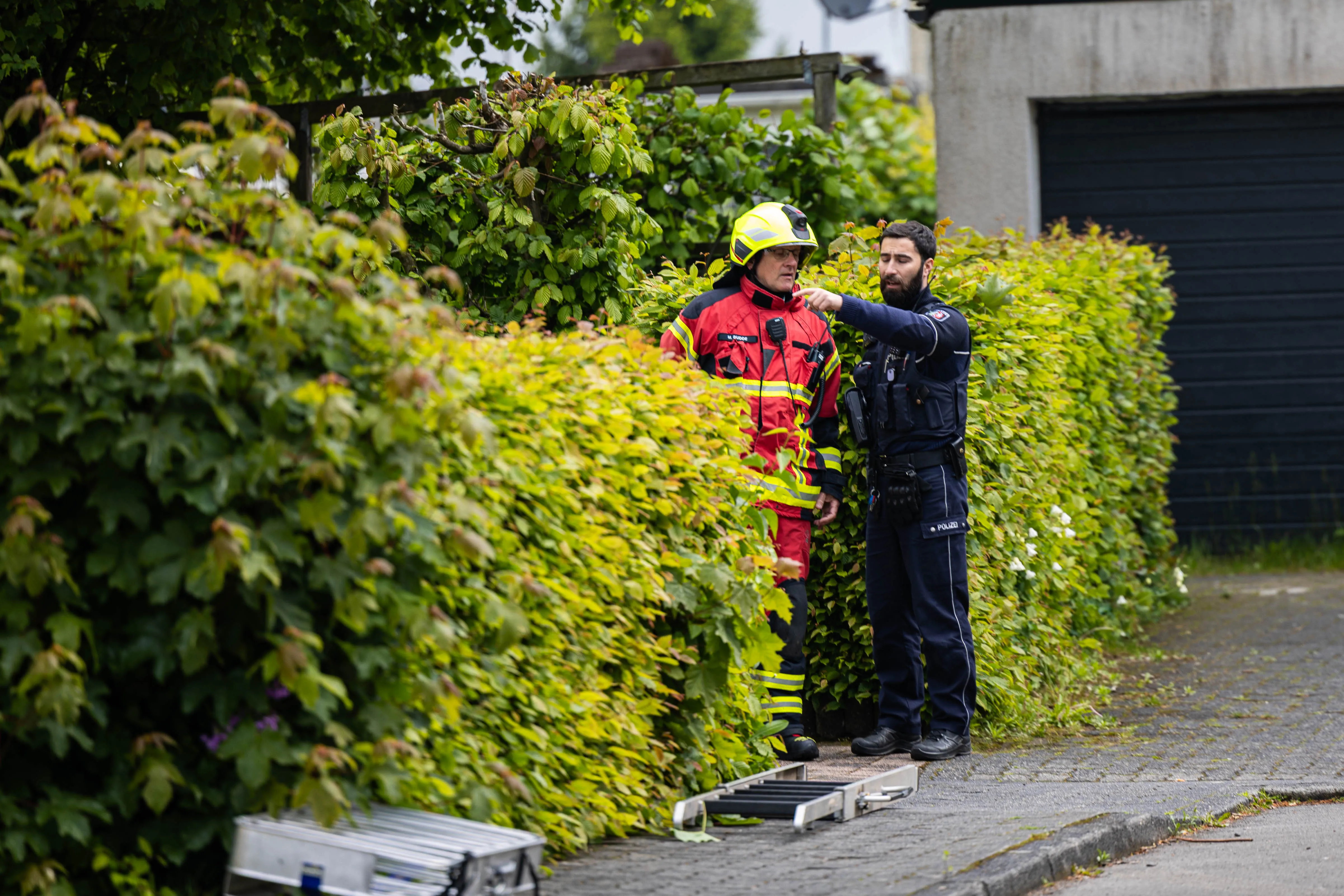 Feuerwehrmann in voller Montur und Polizist besprechen sich neben einer Hecke an einem Einsatzort.