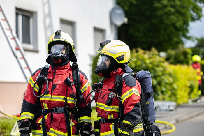 Zwei Feuerwehrleute mit Schutzanzügen und Helmen stehen vor einem Gebäude, im Hintergrund ein weiterer Kollege.