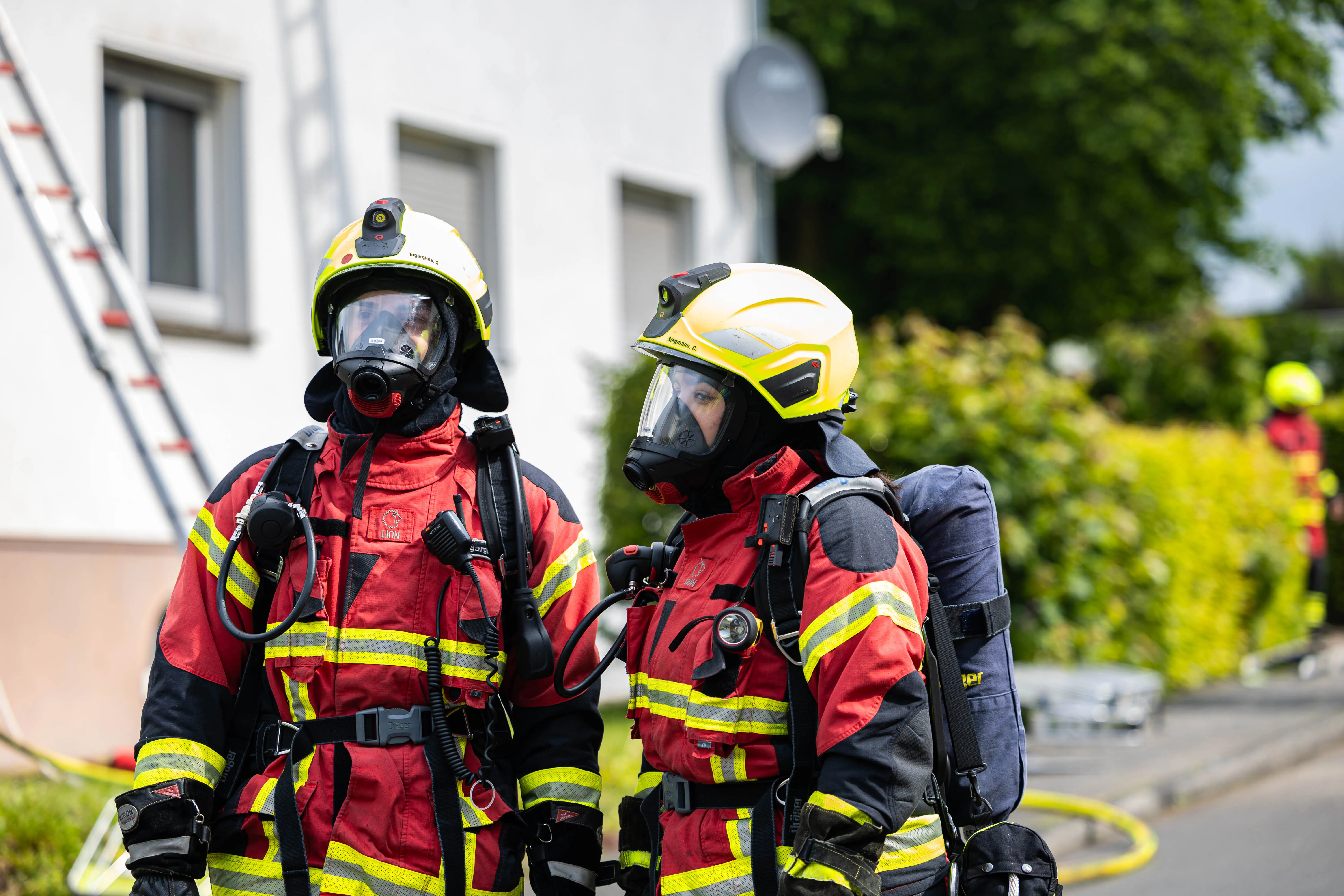 Zwei Feuerwehrleute mit Schutzanzügen und Helmen stehen vor einem Gebäude, im Hintergrund ein weiterer Kollege.