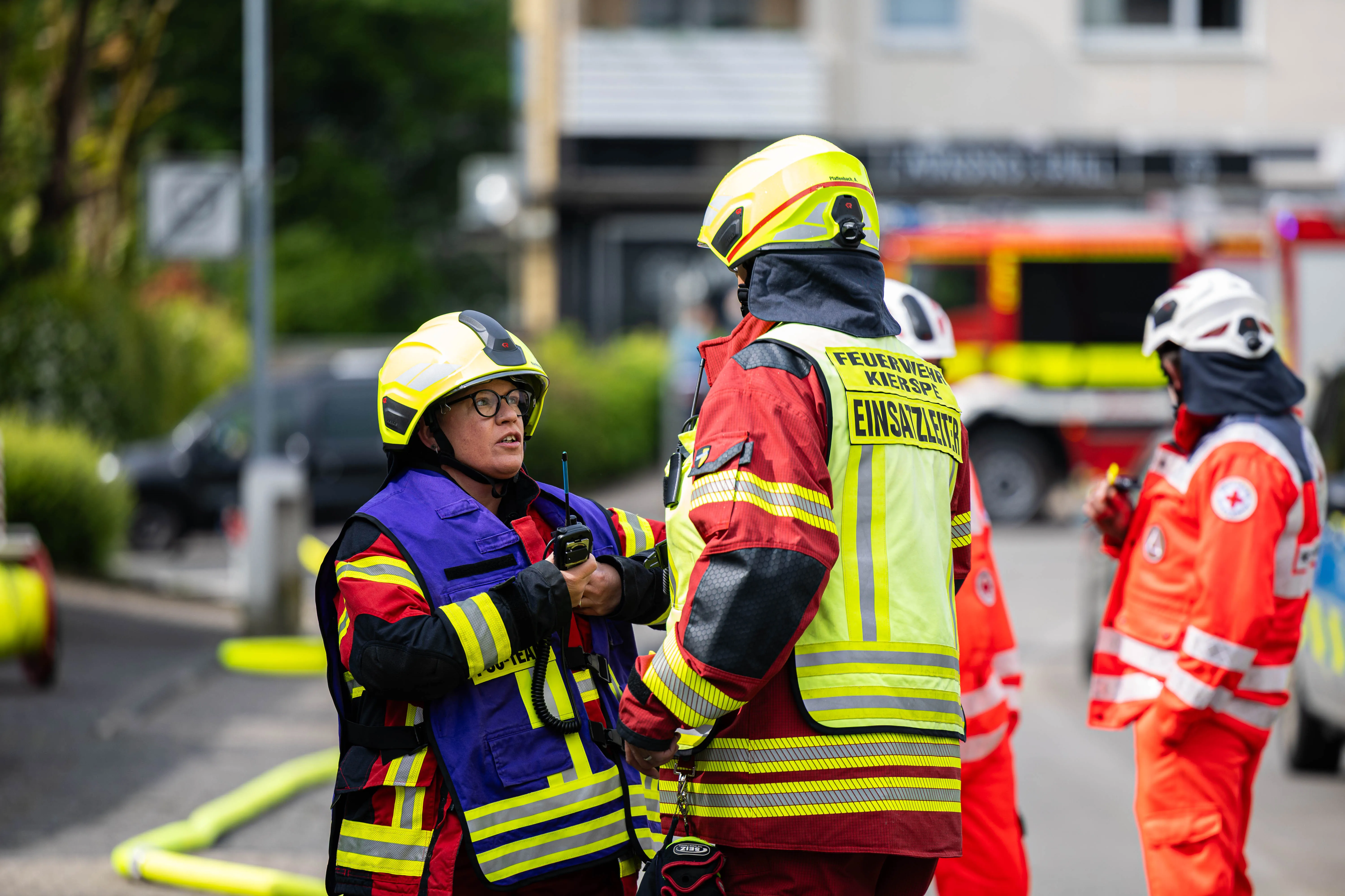 Zwei Feuerwehrleute mit Helmen und Warnwesten besprechen die Lage an einem Einsatzort, im Hintergrund ein weiteres Team und ein rotes Einsatzfahrzeug.