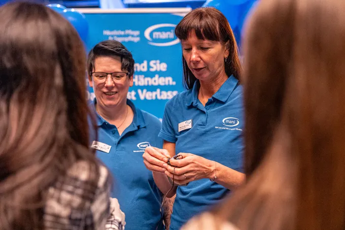 Zwei Frauen in blauen Poloshirts mit dem „mani“-Logo stehen vor einem blauen Banner und interagieren mit Personen im Vordergrund.