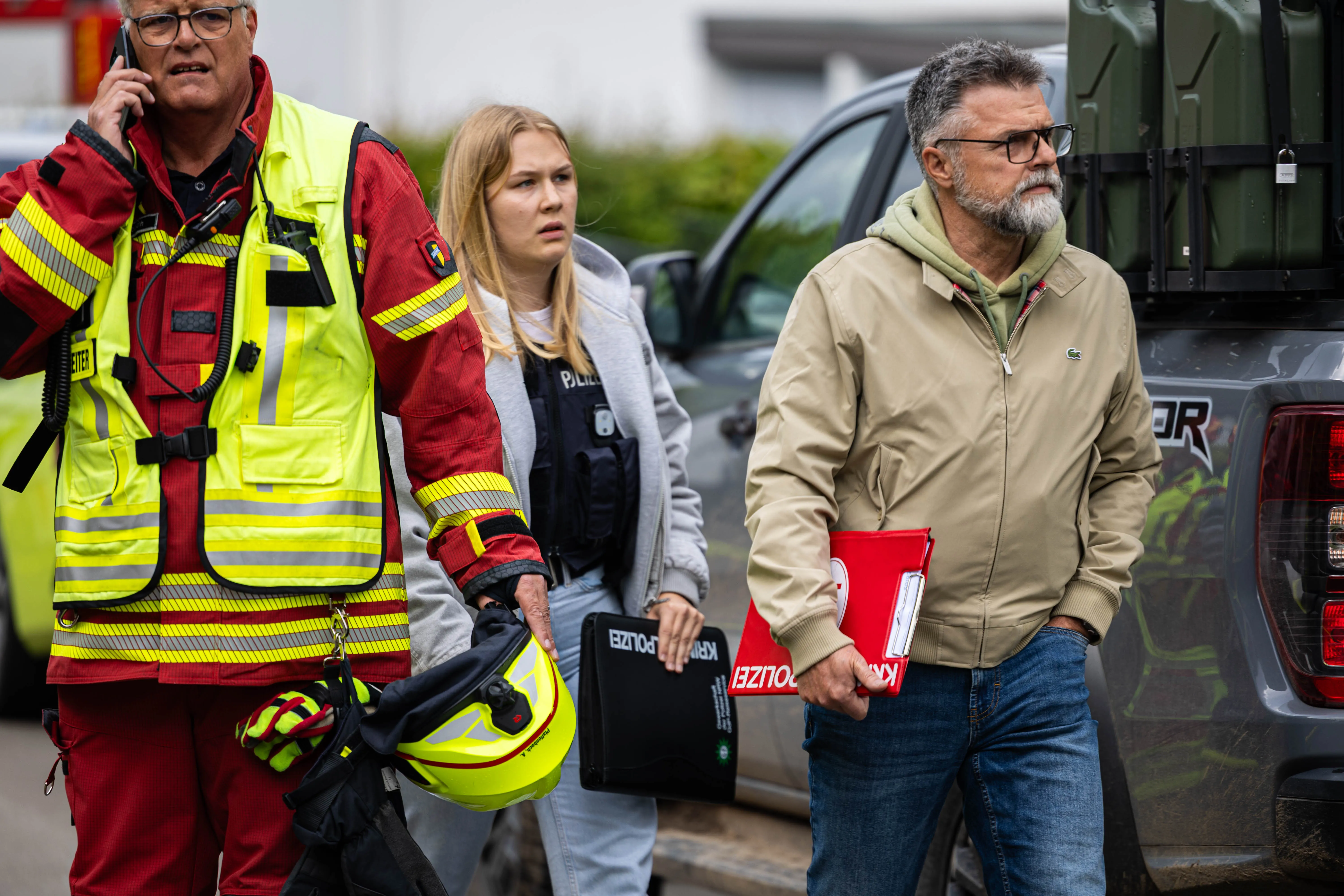 Feuerwehrmann telefoniert während eine Polizistin und ein Mann mit Aktenmappe im Hintergrund stehen, Einsatzfahrzeug im Hintergrund.