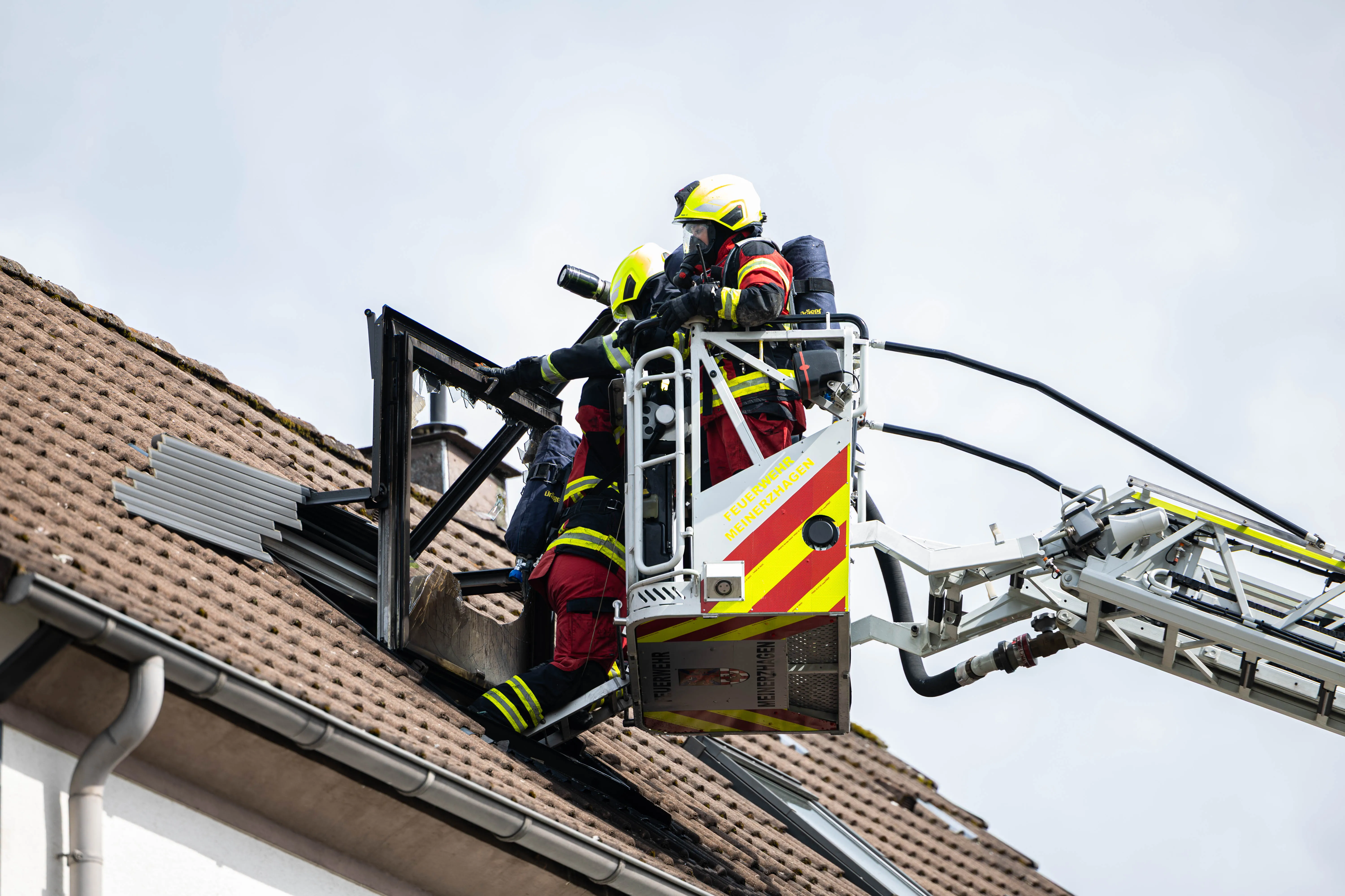 Zwei Feuerwehrleute in voller Montur arbeiten auf einer Hebebühne vor einem roten Ziegeldach. Die Bühne ist mit der Aufschrift „Feuerwehr Meinerzhagen“ versehen.