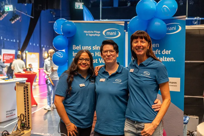 Drei lächelnde Frauen in blauen Polo-Shirts mit „mani“-Logo stehen vor einem blauen Werbebanner und blauen Luftballons.