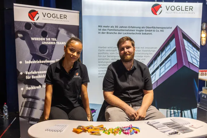 Eine Frau und ein Mann sitzen an einem Messestand vor einem Banner der Firma Vogler Industrielackierungen; auf dem Tisch vor ihnen liegen verschiedene Materialproben.