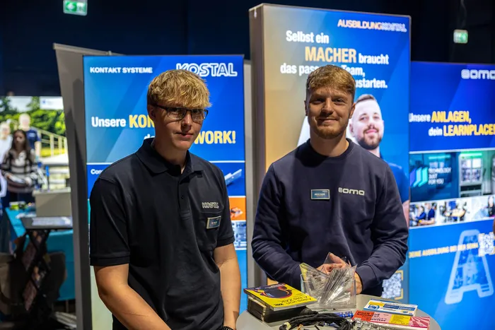 Zwei junge Männer stehen an einem Messestand mit Werbebannern der Firmen Kostal und Soma.