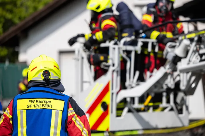 Feuerwehrmann mit Helm und Einsatzjacke vor einem Feuerwehrauto, im Hintergrund weitere Einsatzkräfte vor einem Gebäude.