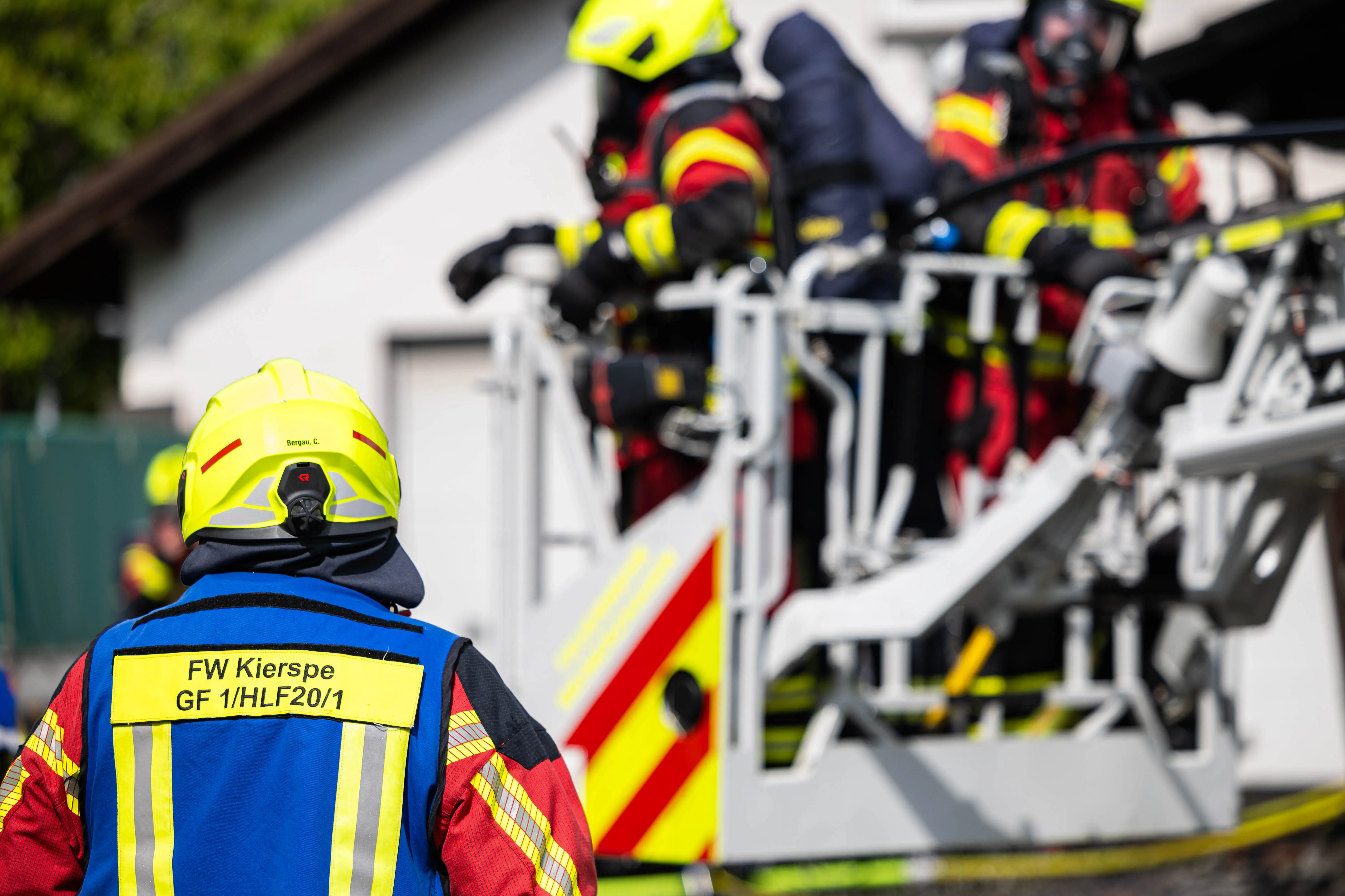 Feuerwehrmann mit Helm und Einsatzjacke vor einem Feuerwehrauto, im Hintergrund weitere Einsatzkräfte vor einem Gebäude.