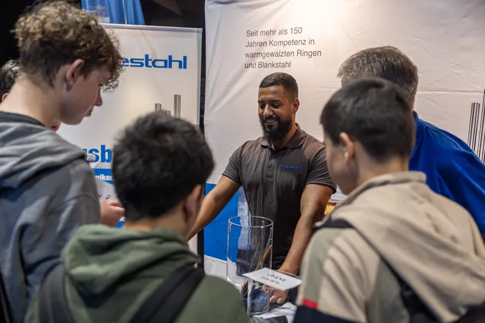 Ein Mann in einem dunklen Poloshirt spricht mit einer Gruppe junger Menschen an einem Messestand der Firma Prostahl.