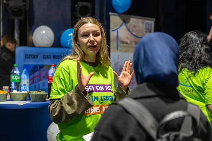 Eine junge Frau in einem grünen T-Shirt mit der Aufschrift „SEI EIN HELFER IM LEBEN EINES KINDES“ spricht gestikulierend mit einer Person, die einen blauen Hidschab trägt.