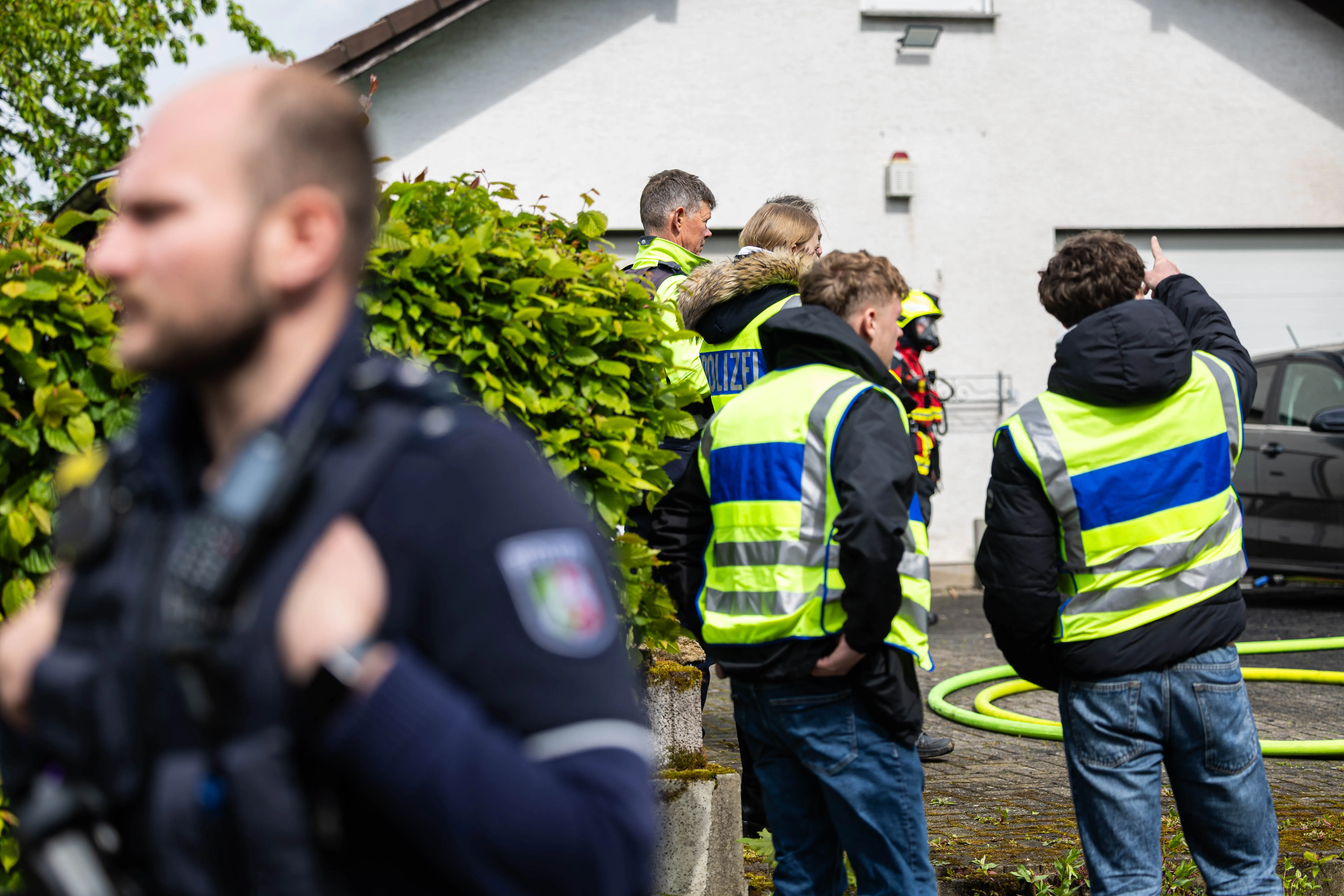 Polizisten und Feuerwehrleute stehen vor einem Wohnhaus, während ein Schlauch am Boden liegt. Im Hintergrund ist ein gepflegter Garten zu sehen.