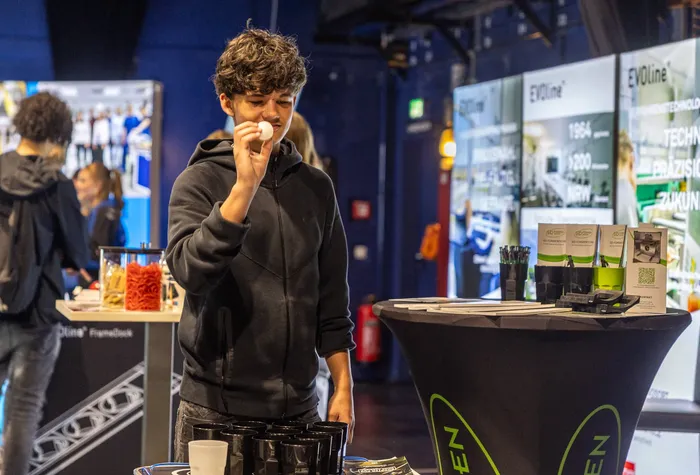 Ein junger Mann in einem dunklen Kapuzenpullover hält einen kleinen weißen Gegenstand vor sein Gesicht an einem Stand der ZAK-Ausbildungsmesse.