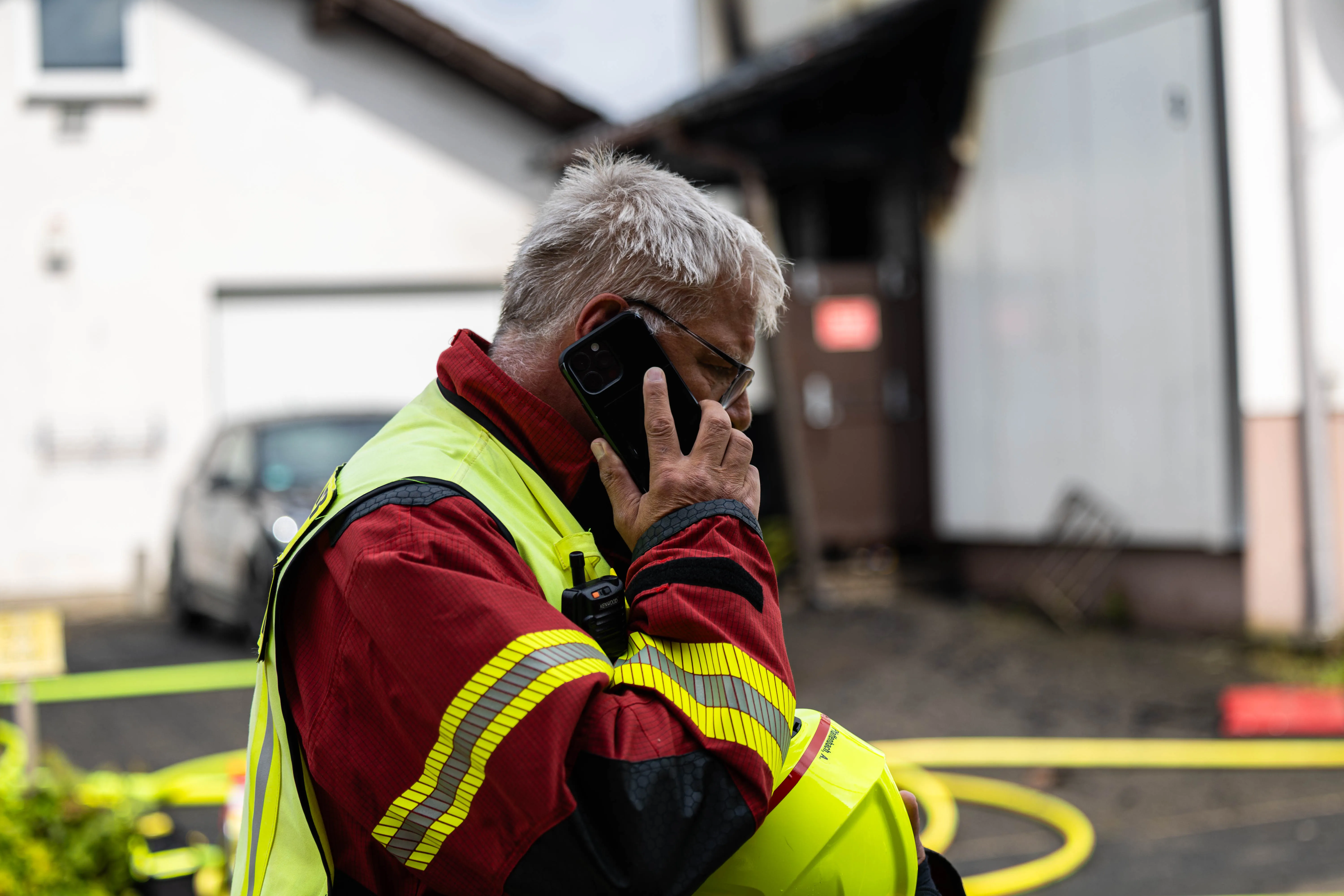 Ein Feuerwehrmann mit grauen Haaren spricht in ein Mobiltelefon, während er eine Warnweste und eine Funkgeräteträger trägt. Im Hintergrund sind Häuser und ein Feuerwehrauto zu sehen.