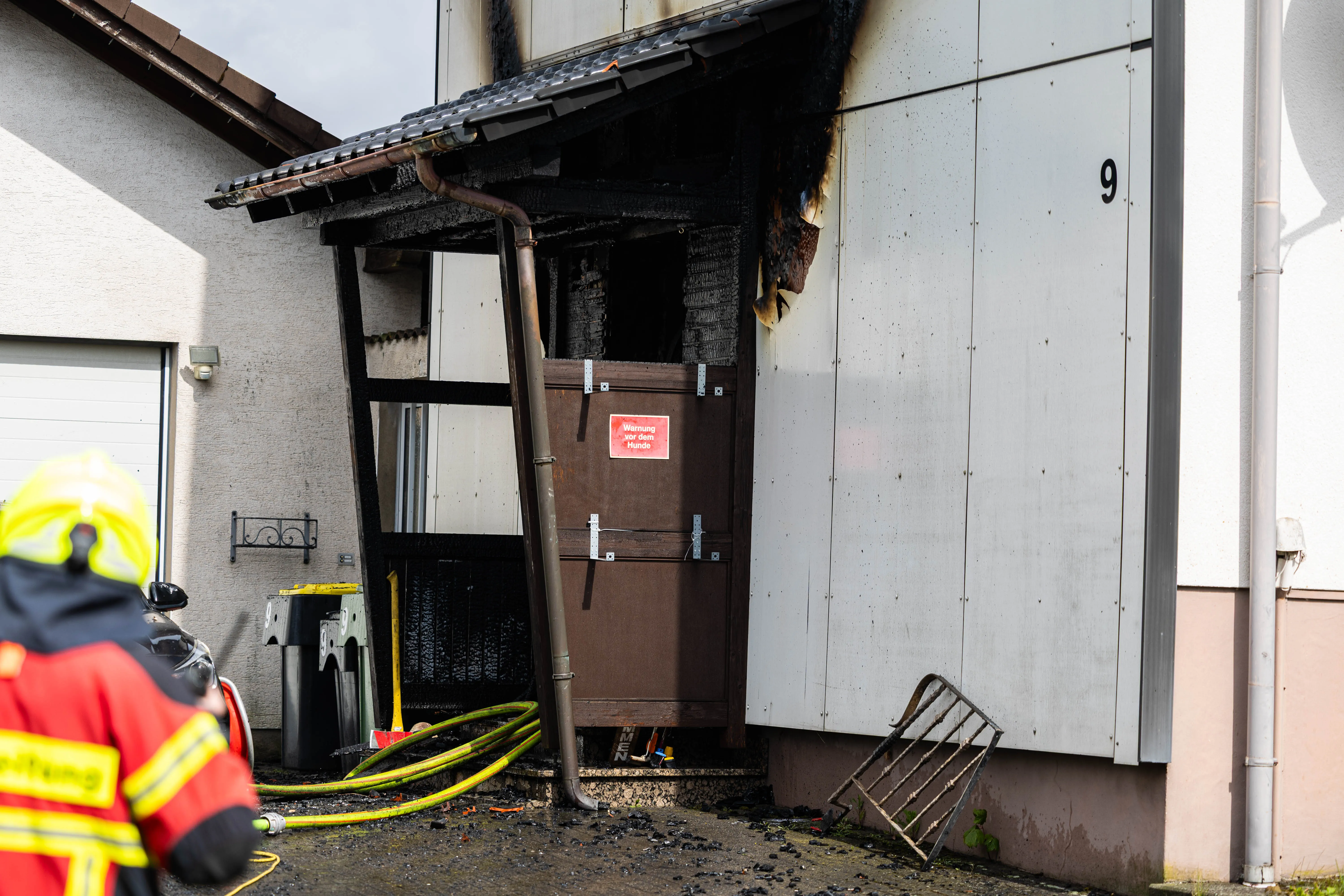 Feuerwehrleute vor einem beschädigten Hauseingang mit Rußspuren nach einem Brand. Ein Schild warnt vor dem Betreten des Bereichs.