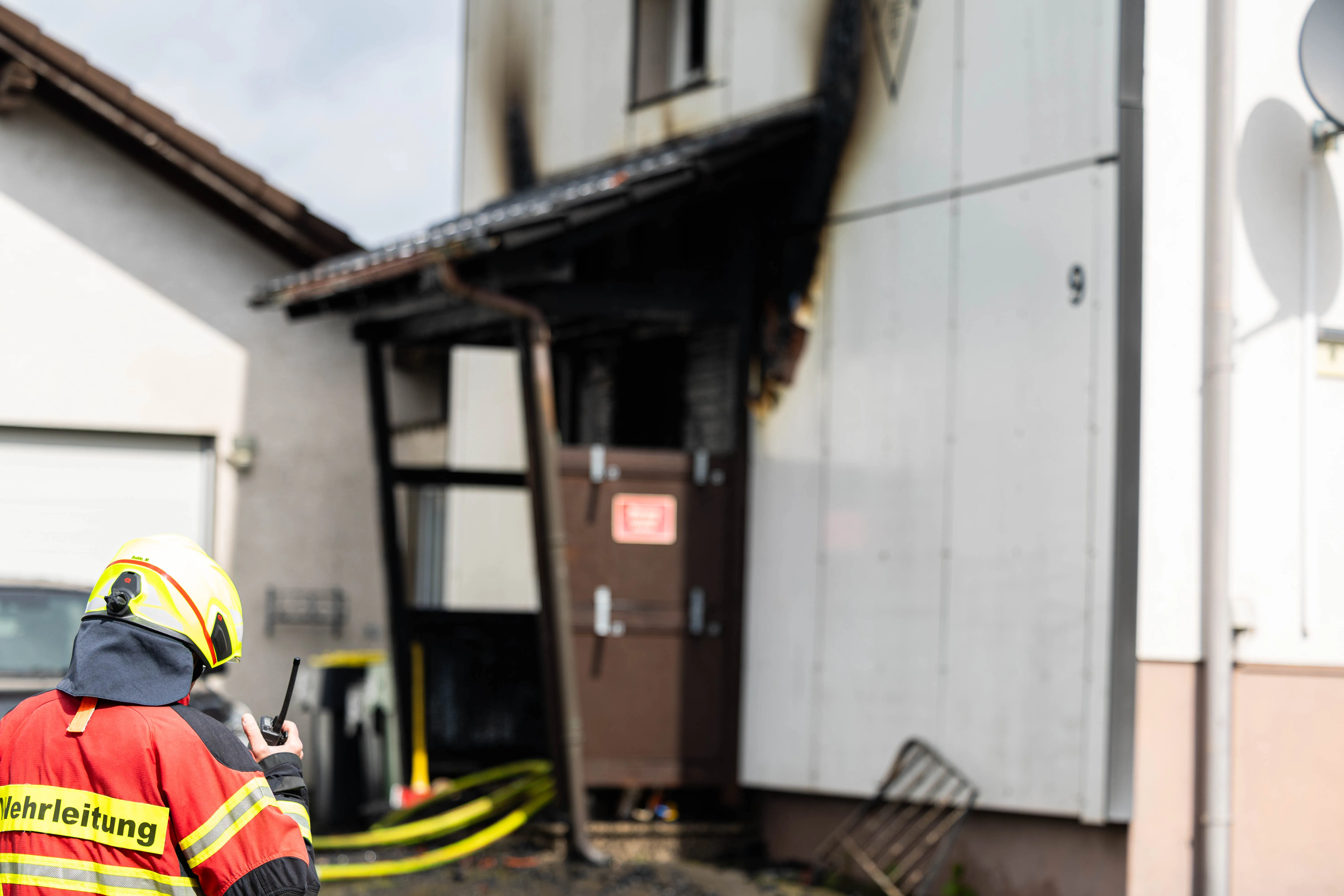 Feuerwehrleute stehen vor einem Haus, dessen Fassade durch einen Brand geschwärzt wurde. Im Vordergrund ist ein Feuerwehrmann mit Funkgerät zu sehen.
