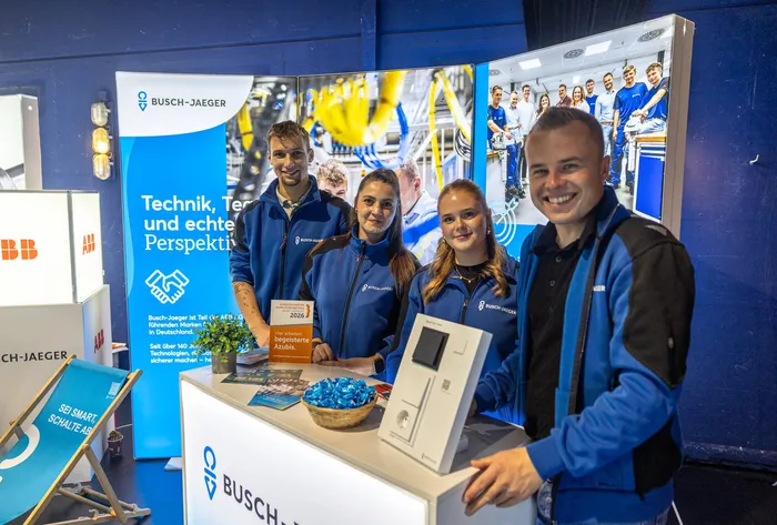 Vier lächelnde junge Menschen in blauen Busch-Jaeger-Jacken stehen hinter einem Infostand.