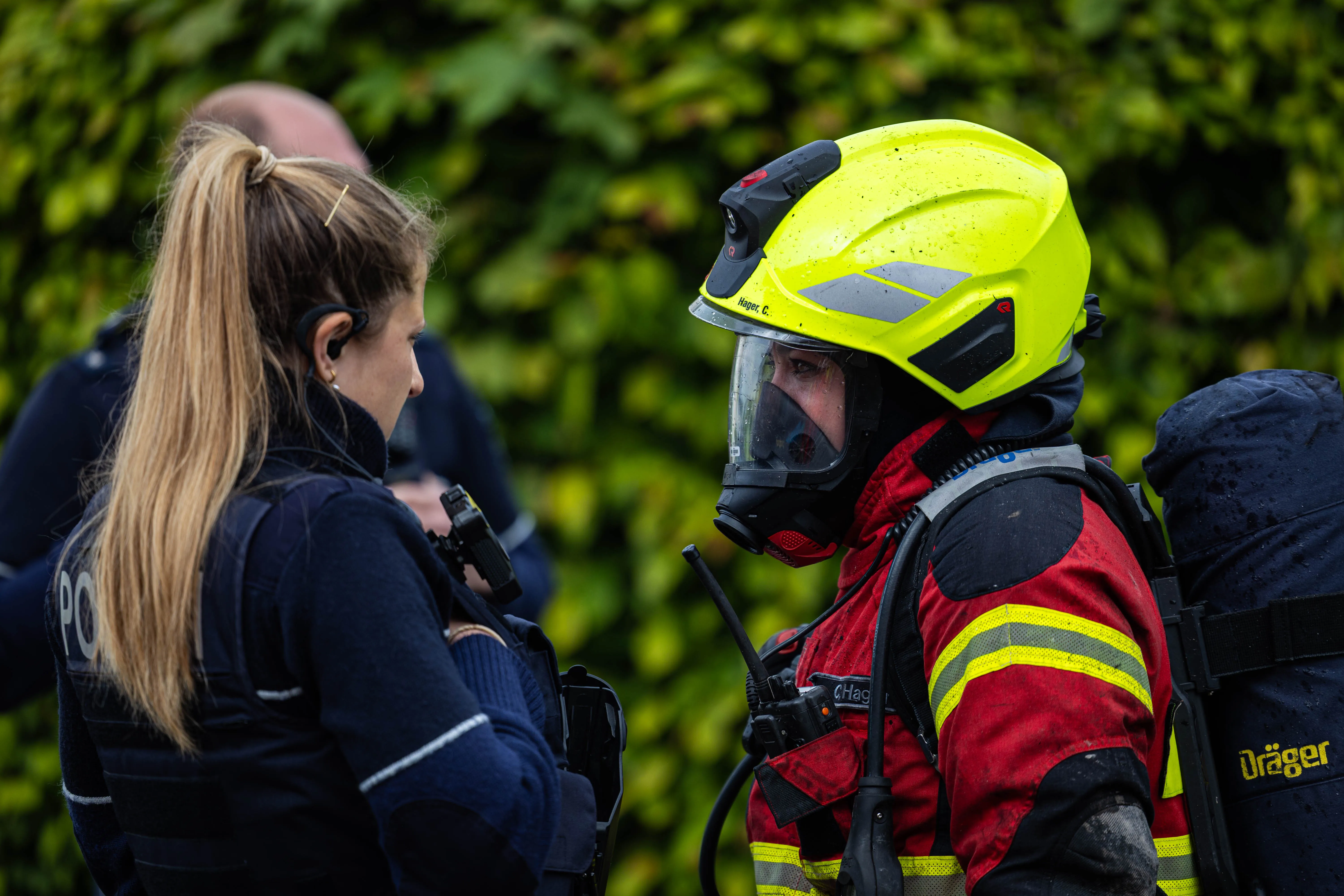 Feuerwehrmann mit Helm und Atemschutzgerät spricht mit einer Polizistin vor grünem Hintergrund während eines Einsatzes in der Gartenstraße.