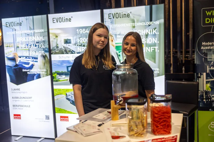 Zwei junge Frauen in schwarzen EVOline-T-Shirts stehen hinter einem Tisch mit Gläsern und Broschüren an einem Messestand.