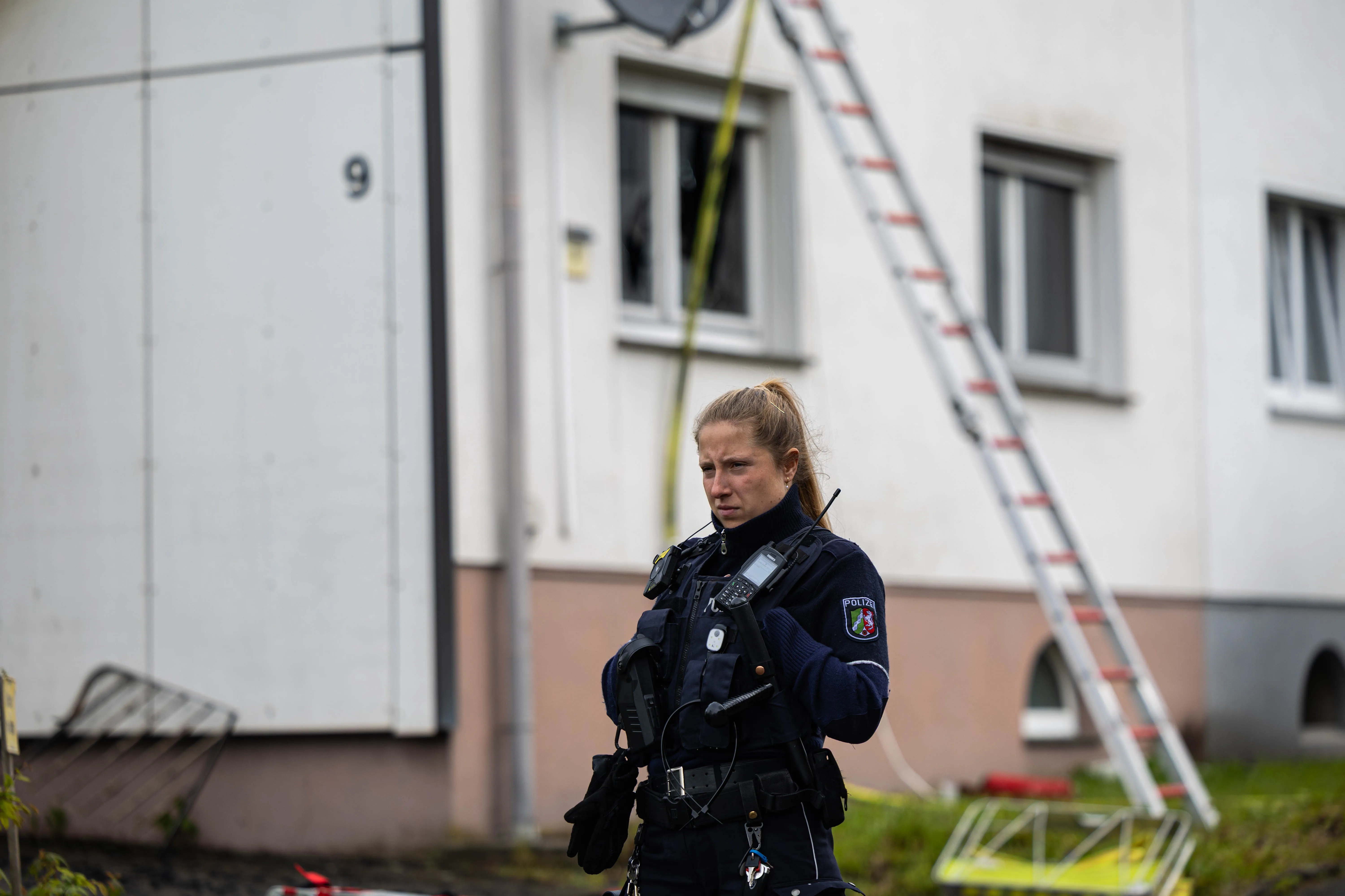 Eine Polizistin steht vor einem zweigeschossigen Wohnhaus mit Brandspuren an einem Fenster. Im Hintergrund ist eine Leiter an der Fassade angelehnt.