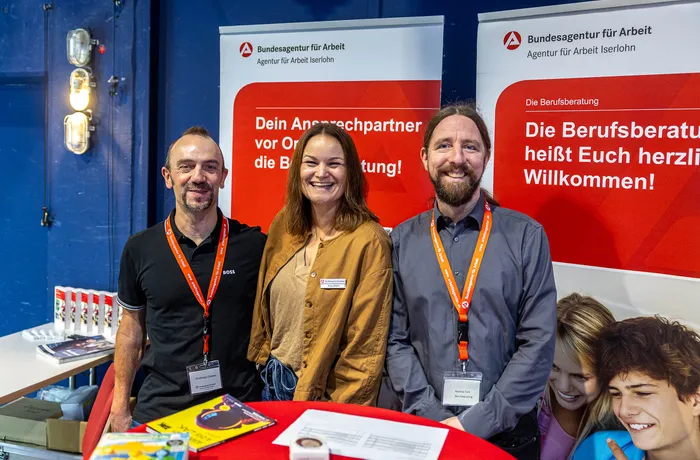 Drei lächelnde Personen stehen hinter einem Infostand der Agentur für Arbeit Iserlohn mit Bannern zur Berufsberatung.