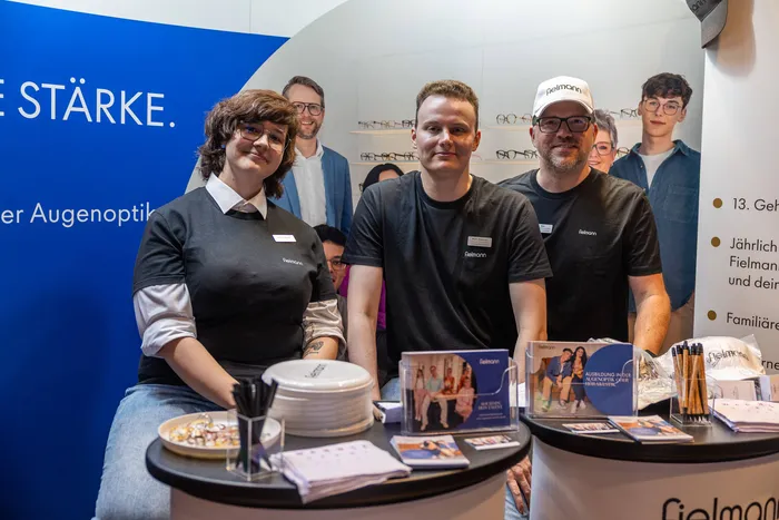 Eine Gruppe von Menschen, teilweise in schwarzen T-Shirts mit Fielmann-Logo, steht hinter einem Stand mit Werbematerialien.